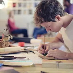 Młody mężczyzna w białej koszulce rysuje precyzyjny plan na desce kreślarskiej przy użyciu ołówka i linijki, w tle widoczne osoby i akcesoria biurowe pod lampą.