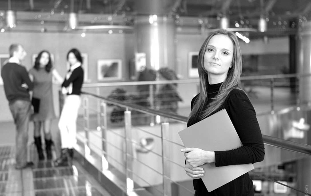 Czarno-białe zdjęcie kobiety trzymającej teczkę na tle nowoczesnego biura z rozmawiającymi osobami w tle.