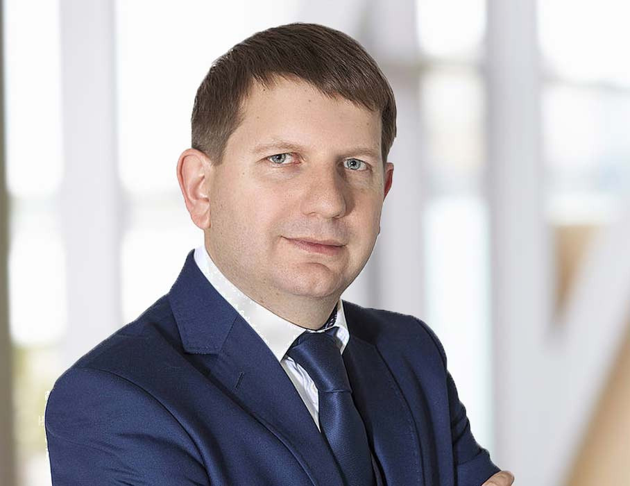 Portret mężczyzny w granatowym garniturze i krawacie, z rękami założonymi na piersiach, na rozmytym tle biurowym.