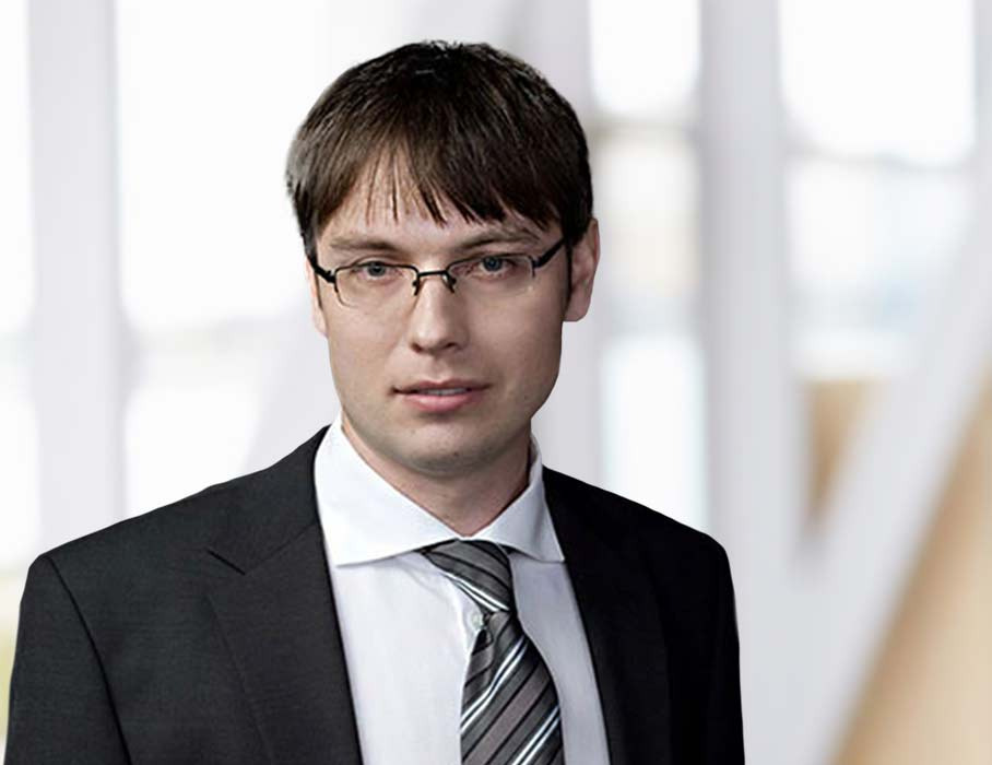 Portret adwokata w okularach, w czarnym garniturze i krawacie w szaro-białe paski, na rozmytym tle biura.