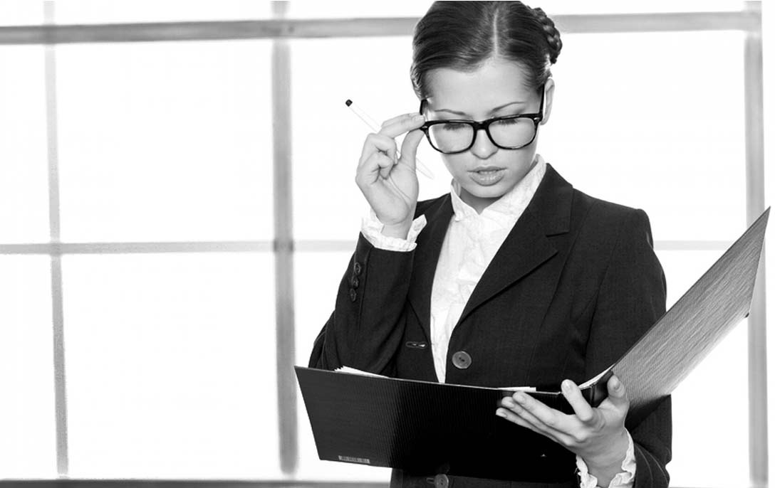 Czarno-białe zdjęcie kobiety w okularach i garniturze, trzymającej czarny segregator i poprawiającej okulary długopisem, na tle okna.