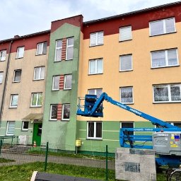 Wysięgnik podnośnika koszowego przy bloku z widocznymi śladami zabrudzeń na elewacji, sugerujący etap czyszczenia. Część elewacji jest wyraźnie czystsza.