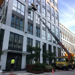 Pracownik w żółtej kamizelce czyści dolne okna budynku biurowego za pomocą długiej lancy, podczas gdy drugi pracownik czyści okna na wysokości za pomocą podnośnika koszowego.