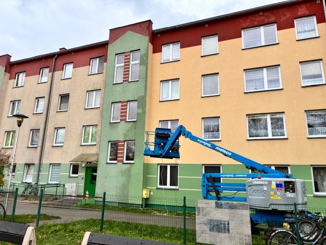 Wysięgnik podnośnika koszowego przy bloku z widocznymi śladami zabrudzeń na elewacji, sugerujący etap czyszczenia. Część elewacji jest wyraźnie czystsza.