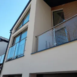 Nowoczesny dom z jasną elewacją, szklanymi balustradami balkonów i czarnymi ramami okiennymi, widziany z dołu pod kątem.