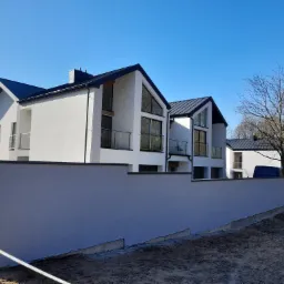 Nowoczesne, dwukondygnacyjne domy w zabudowie szeregowej z białymi elewacjami i grafitowymi dachami, widoczne za świeżo otynkowanym, szarym murem oporowym, w słoneczny dzień.