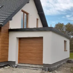 Nowoczesny dom jednorodzinny z elewacją w dwóch odcieniach: jasna, gładka i drewnopodobna, z widoczną bramą garażową o panelowej strukturze i oknami z drewnianą ramą.