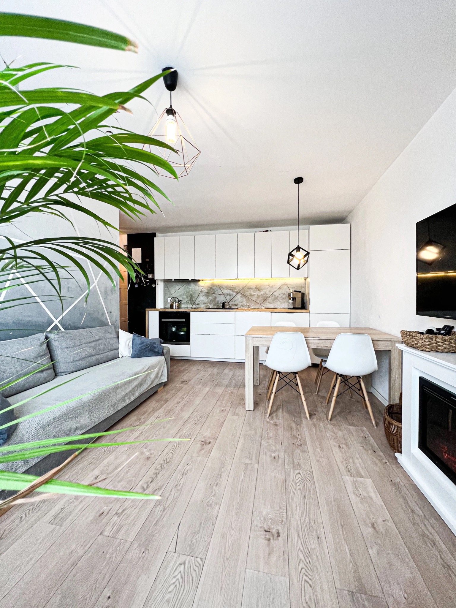 Jasne mieszkanie z drewnianą podłogą, szarą sofą z geometrycznym wzorem na ścianie, białą kuchnią z drewnianym blatem, stołem z białymi krzesłami i dekoracyjnym kominkiem.