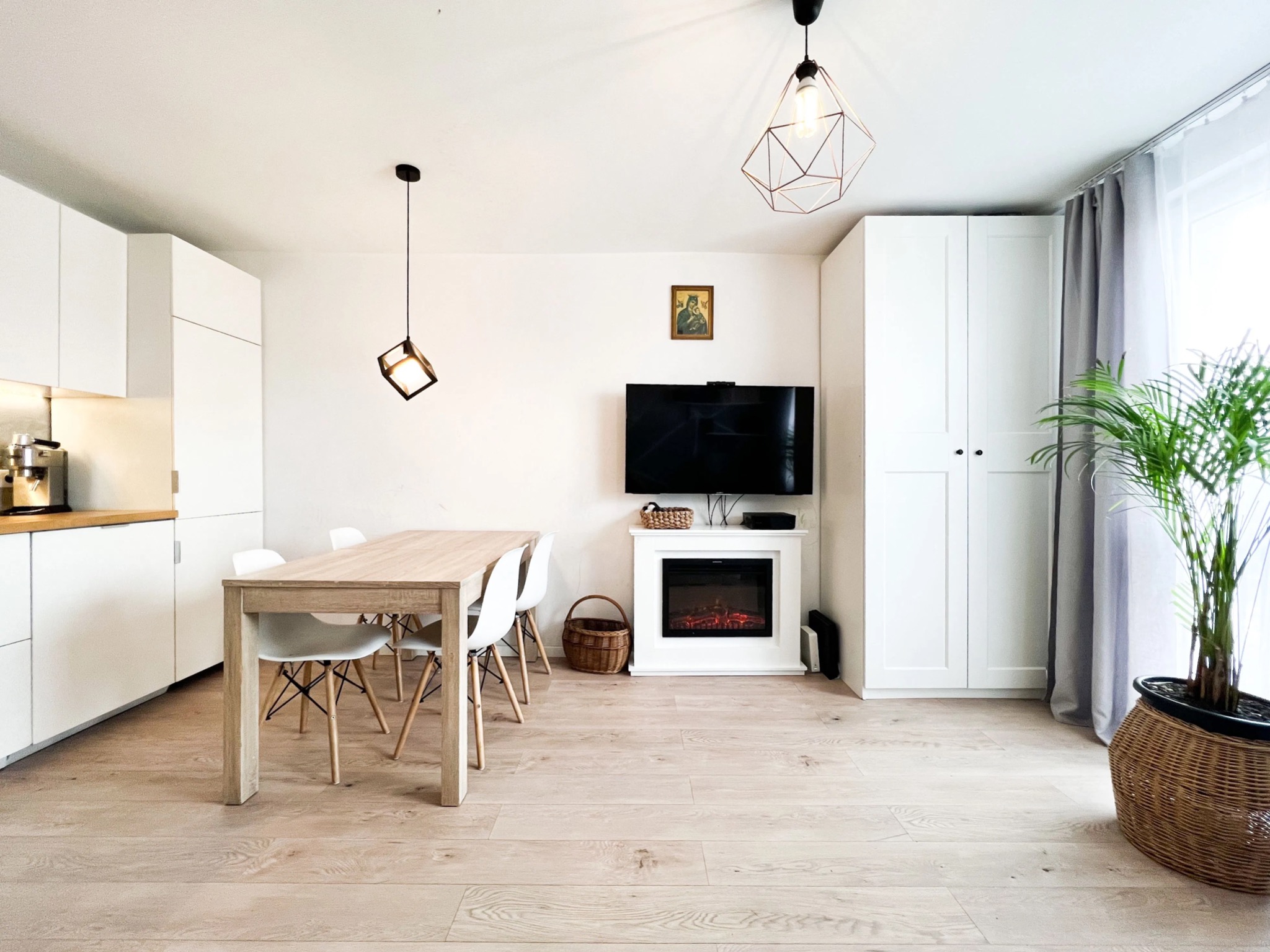 Jasne wnętrze z drewnianym stołem, białymi krzesłami, szafkami i kominkiem elektrycznym, oświetlone geometrycznymi lampami wiszącymi.