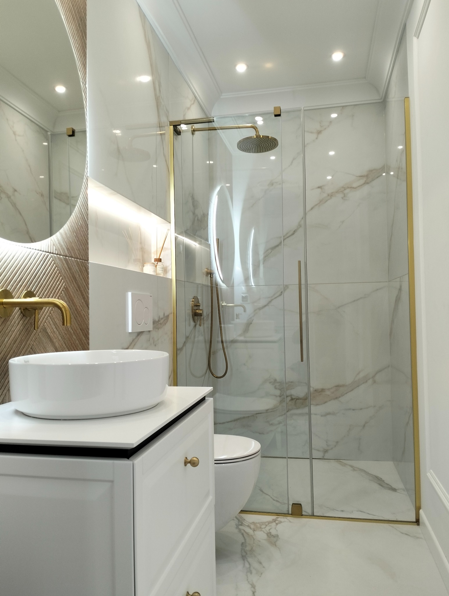 Elegancka łazienka z marmurowymi płytkami, kabiną prysznicową ze złotymi detalami, okrągłym lustrem z podświetleniem i białą umywalką nablatową na szafce.