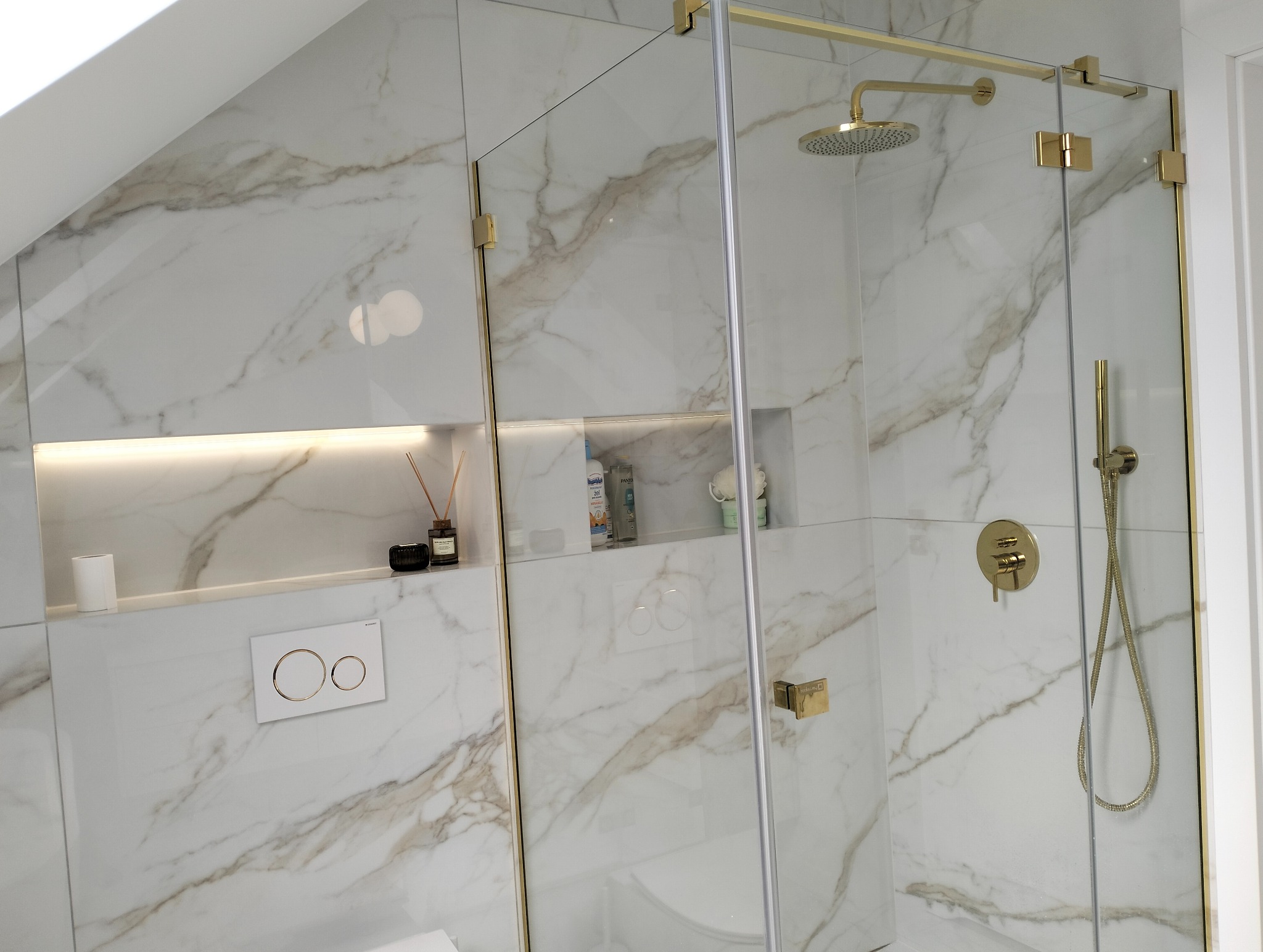 Nowoczesna kabina prysznicowa z białymi płytkami imitującymi marmur, złotymi okuciami i deszczownicą, wnęki z oświetleniem i akcesoriami łazienkowymi.