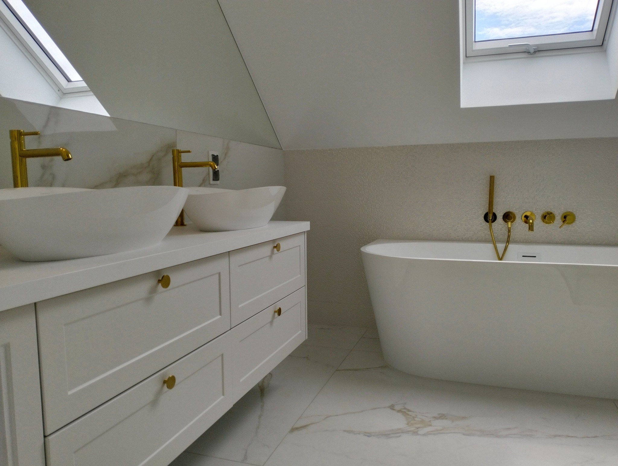 Nowoczesna łazienka na poddaszu z dwoma umywalkami nablatowymi, złotą armaturą i wolnostojącą wanną, wykończona płytkami imitującymi marmur.