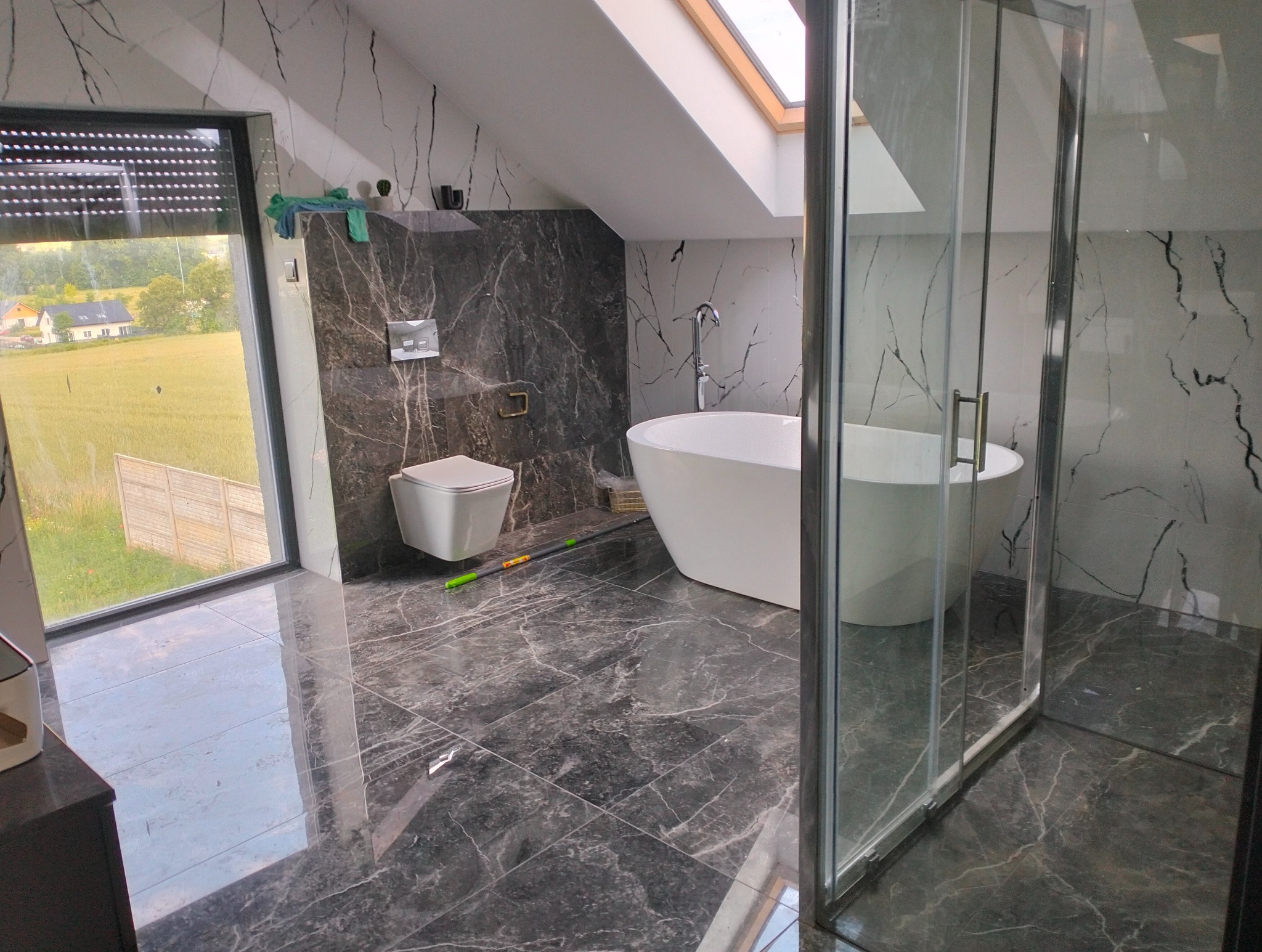 Nowoczesna łazienka na poddaszu z oknem dachowym, wykończona płytkami imitującymi marmur, z wolnostojącą wanną, toaletą podwieszaną i szklaną kabiną prysznicową.