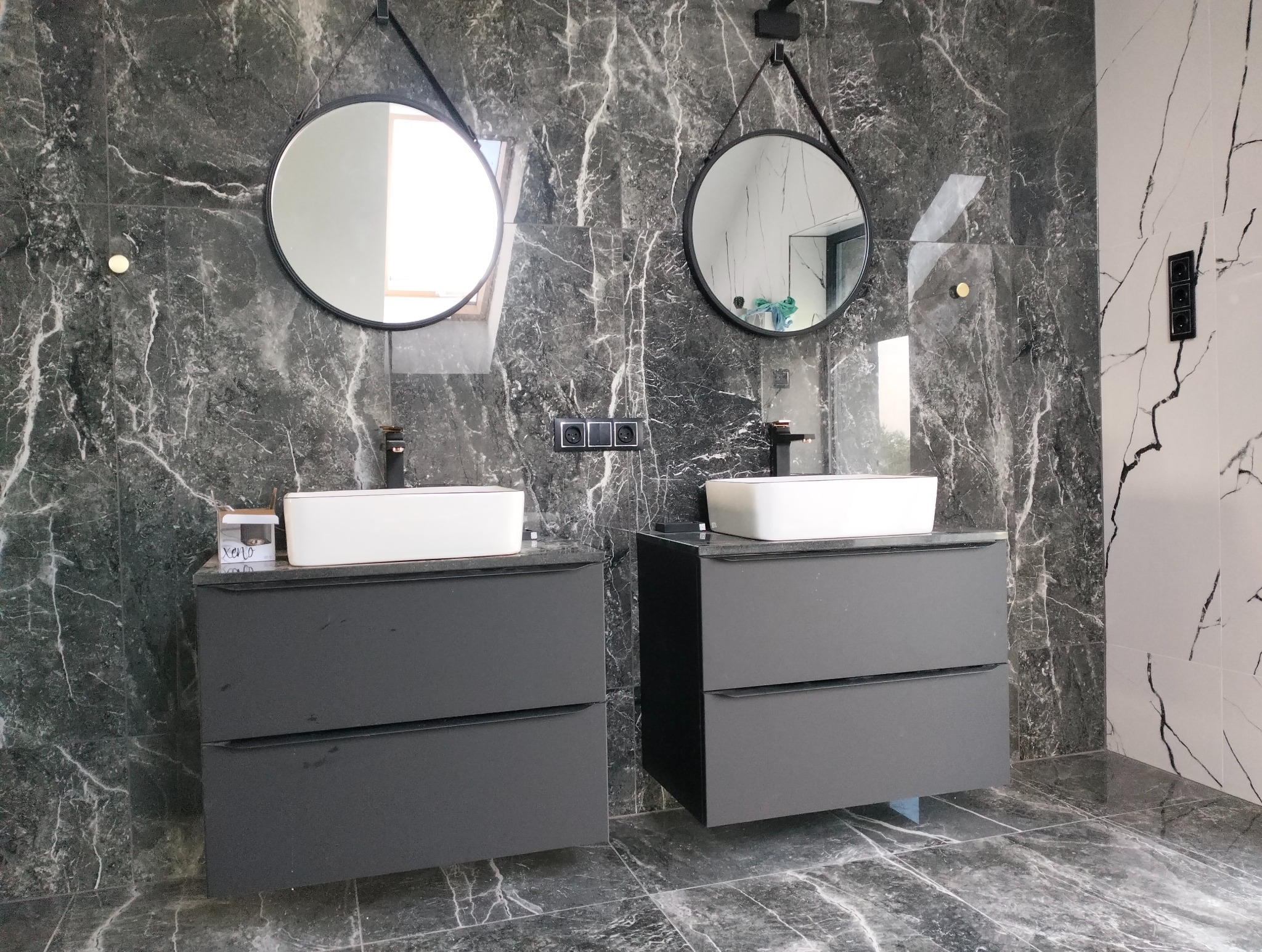 Nowoczesna łazienka z dwoma umywalkami nablatowymi na szarych szafkach, okrągłe lustra na ścianie z płytek imitujących marmur, czarne baterie i gniazdka elektryczne.