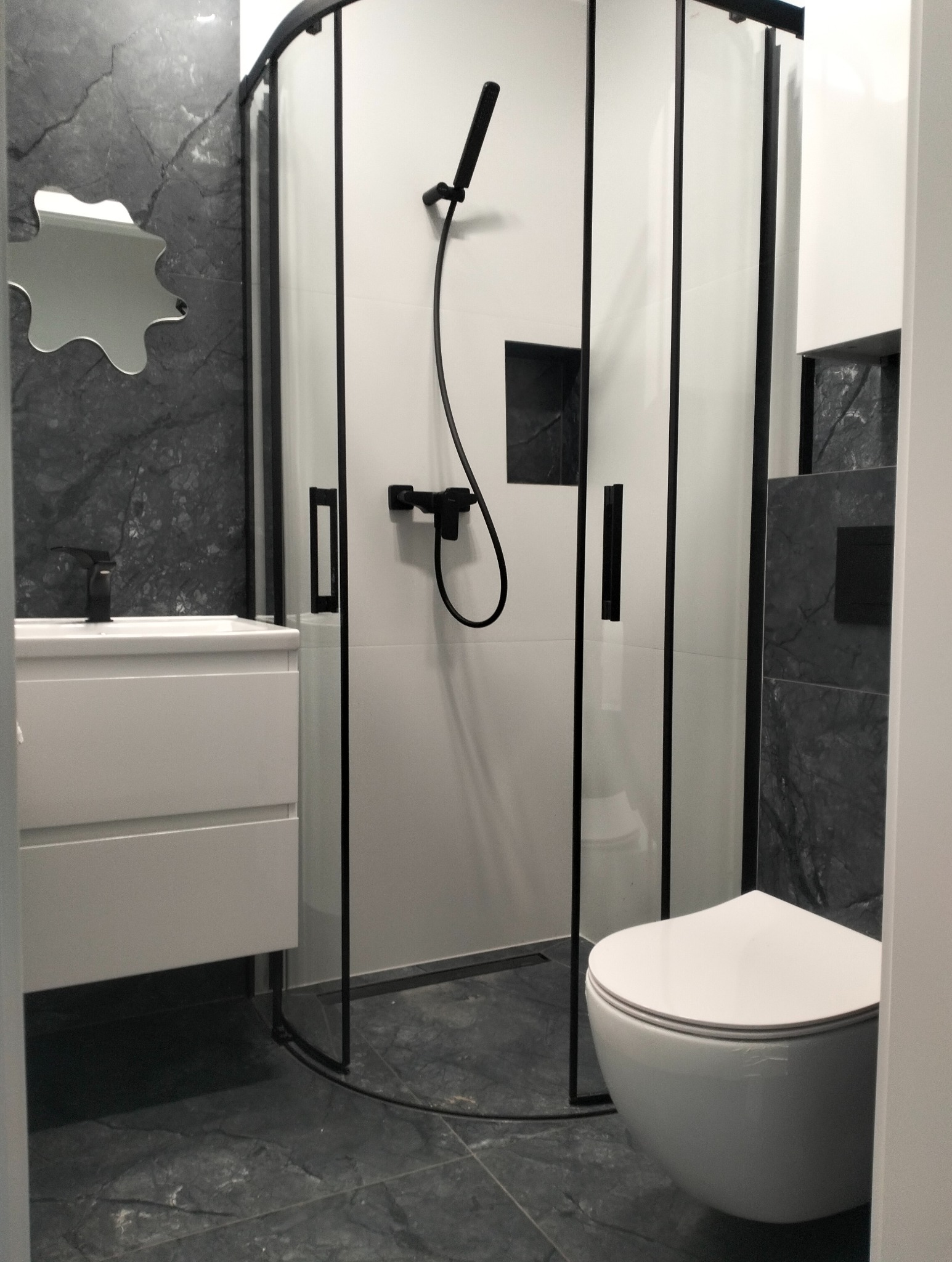 Nowoczesna łazienka z kabiną prysznicową z czarnymi ramami, umywalką z szafką, toaletą podwieszaną i lustrem o nieregularnym kształcie na ścianie wyłożonej szarymi płytkami imitującymi marmur.