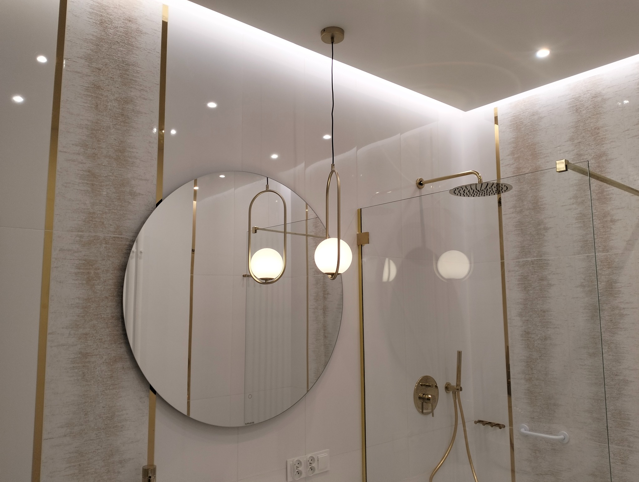 Nowoczesna łazienka z okrągłym lustrem podświetlonym od tyłu, złotymi akcentami na armaturze i płytkach oraz deszczownicą za szklaną ścianką kabiny.