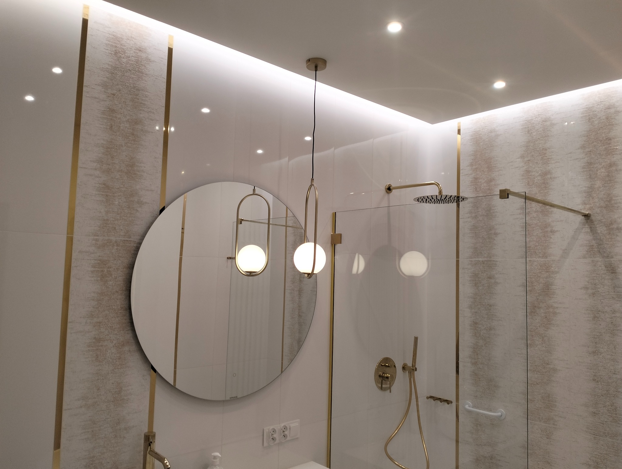 Nowoczesna łazienka z kabiną prysznicową oddzieloną szybą, okrągłym lustrem z podświetleniem i wiszącą lampą z dwoma kulami, wykończona białymi płytkami z akcentami w kolorze złota.