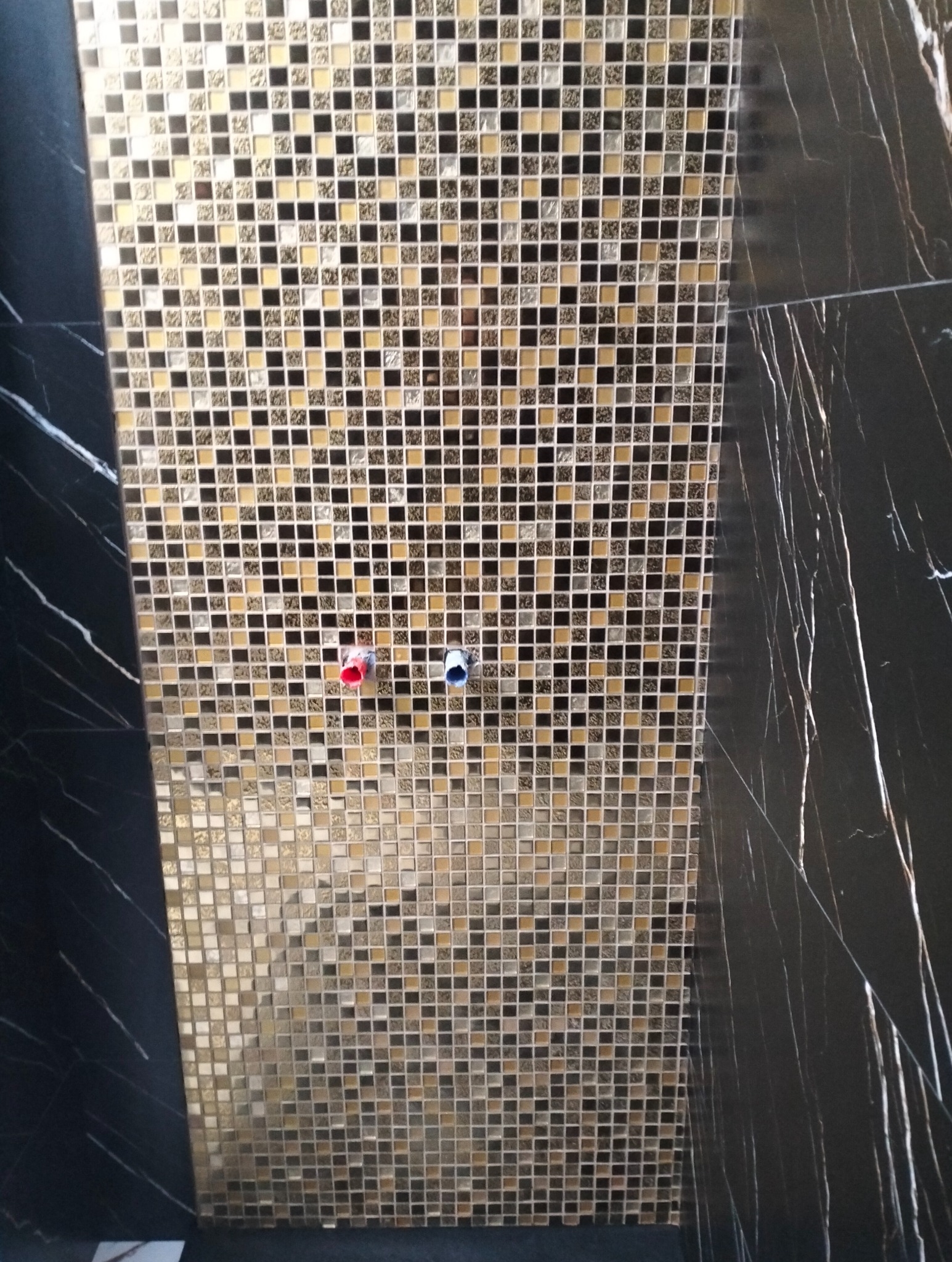 Fragment ściany wyłożonej mozaiką z drobnych, kwadratowych płytek w odcieniach złota, brązu i czerni, z widocznymi przyłączami wodnymi z czerwoną i niebieską zaślepką, graniczącej z czarną płytą...