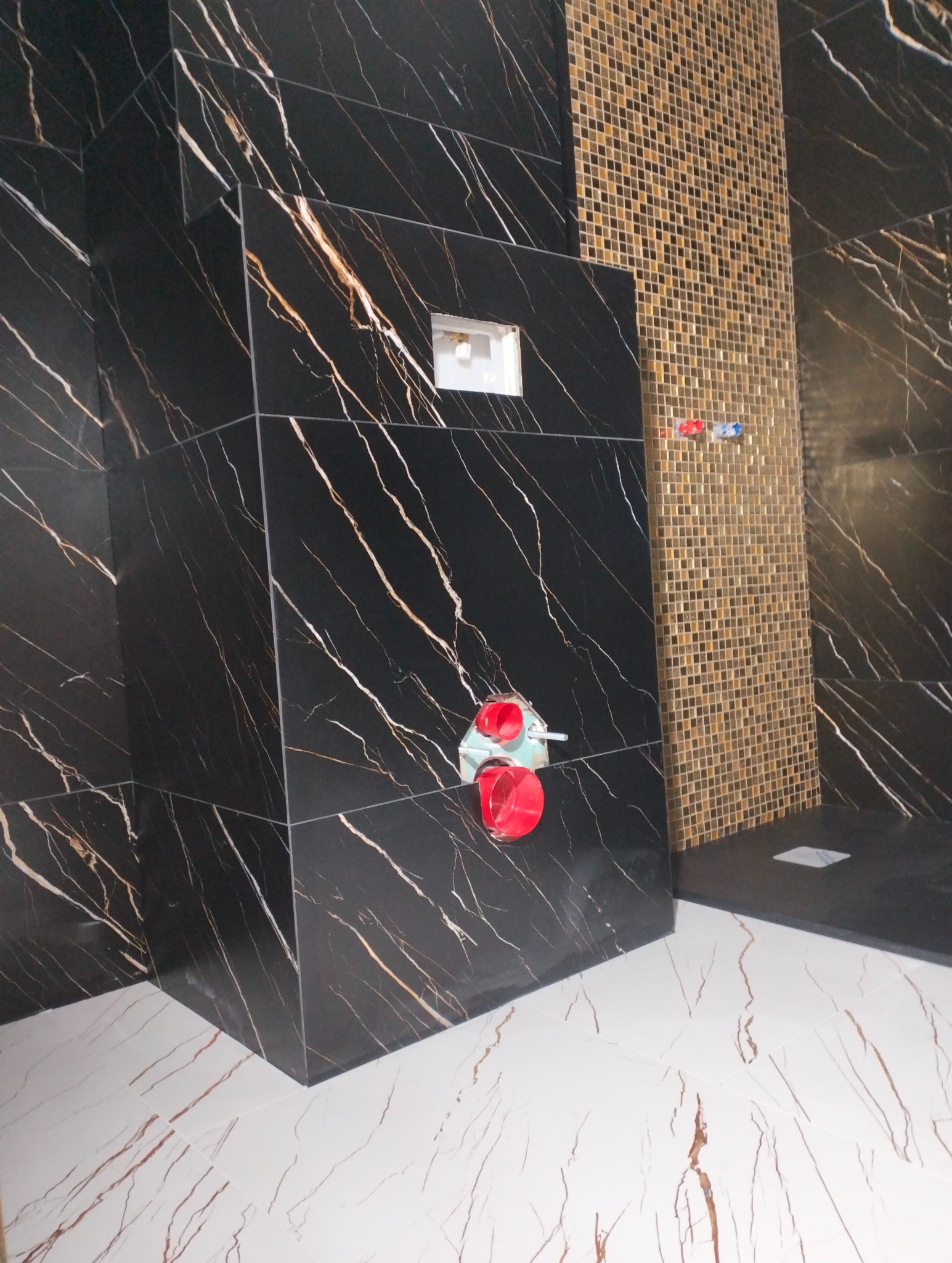 Częściowo wykończona łazienka z czarnymi płytkami imitującymi marmur z białymi żyłami na ścianach i mozaiką w odcieniach brązu w strefie prysznica, widoczne elementy instalacji sanitarnej, białe...