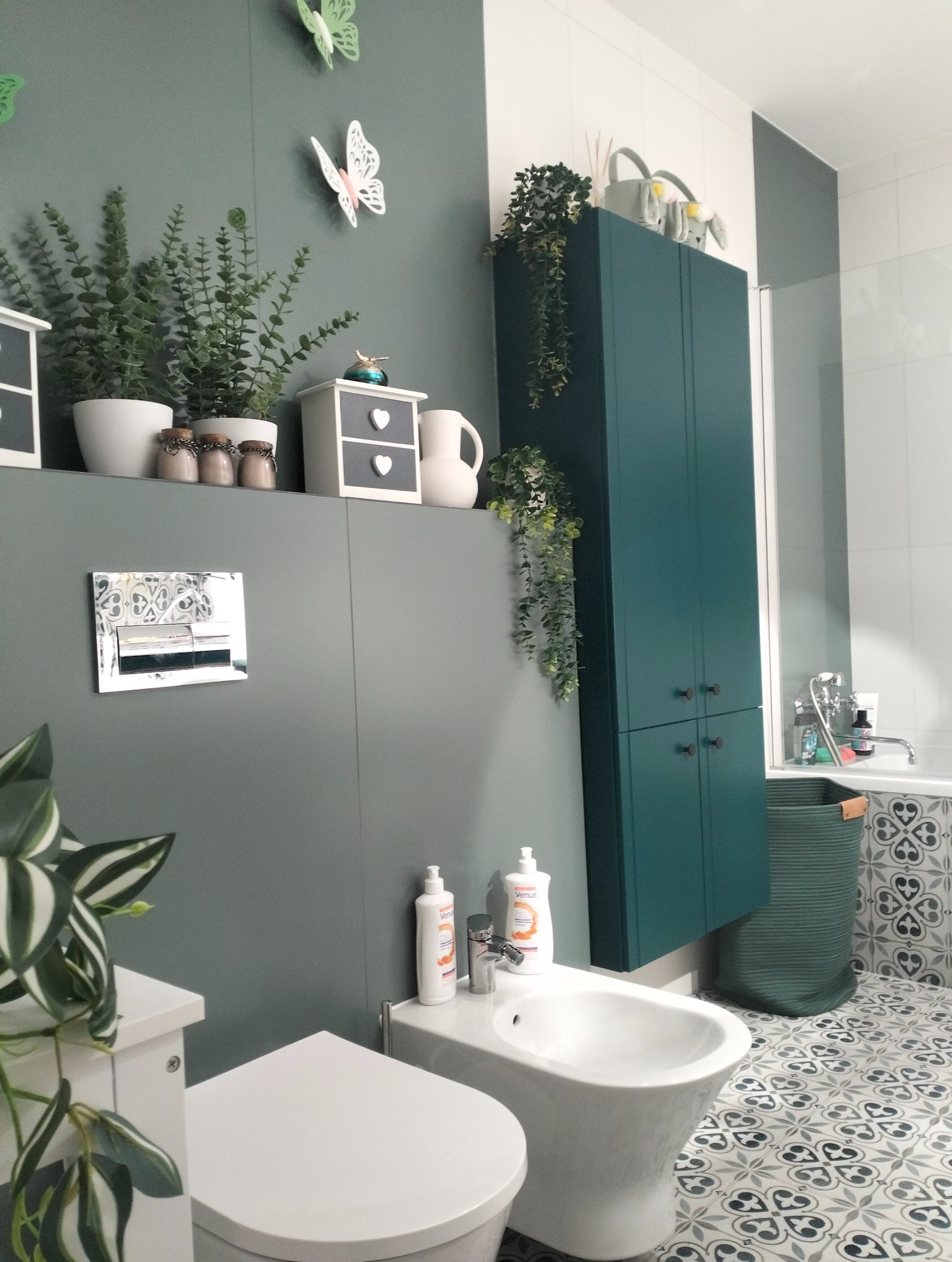 Estetyczna łazienka w Stargardzie z zielonymi ścianami, białymi sanitariatami, wzorzystą podłogą i dekoracyjnymi roślinami, w tym wiszącymi pnączami na szafce.