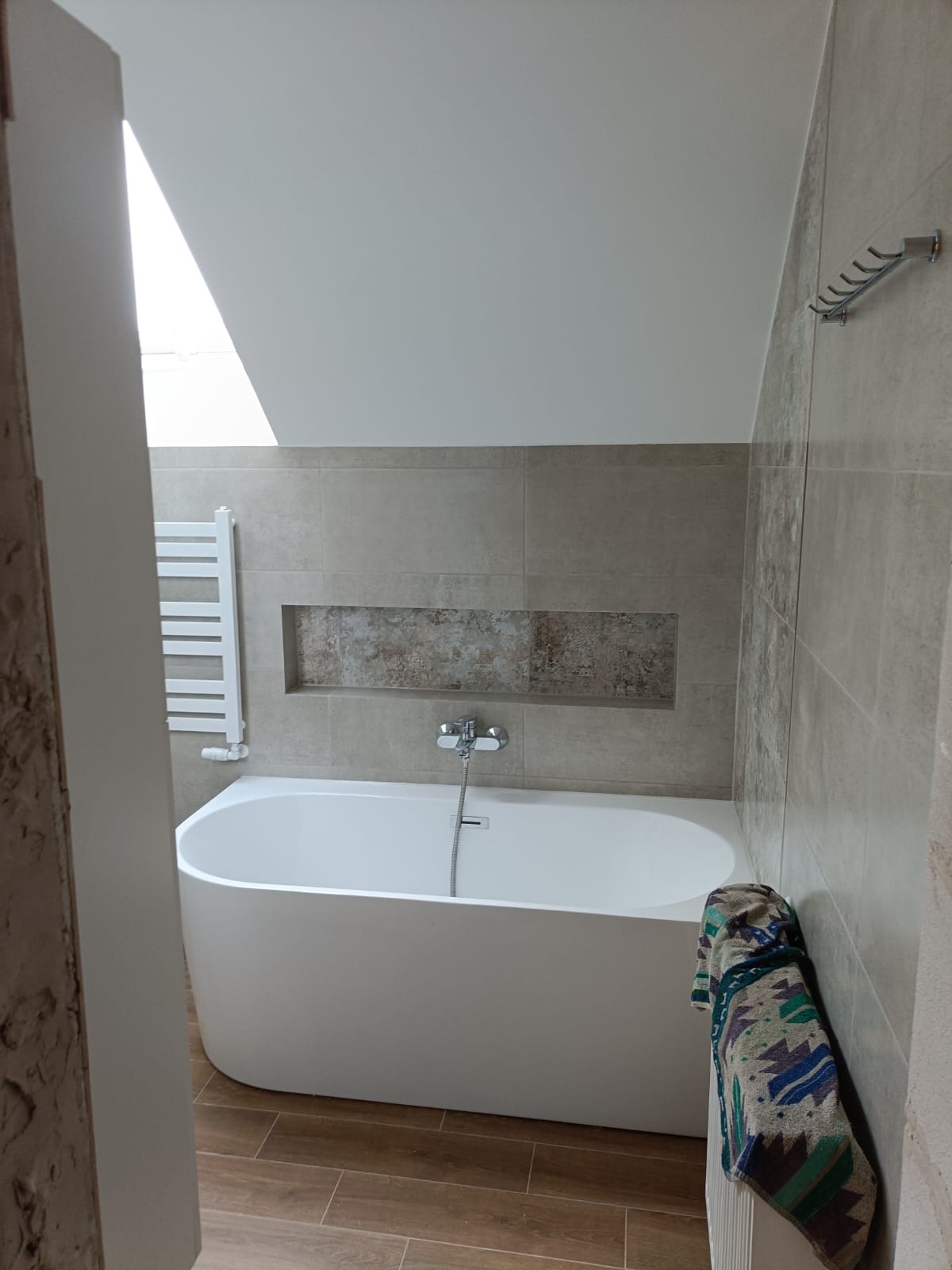 Nowoczesna łazienka na poddaszu z białą wanną wolnostojącą, grzejnikiem drabinkowym, beżowymi płytkami ściennymi z dekoracyjną wnęką i drewnopodobną podłogą.