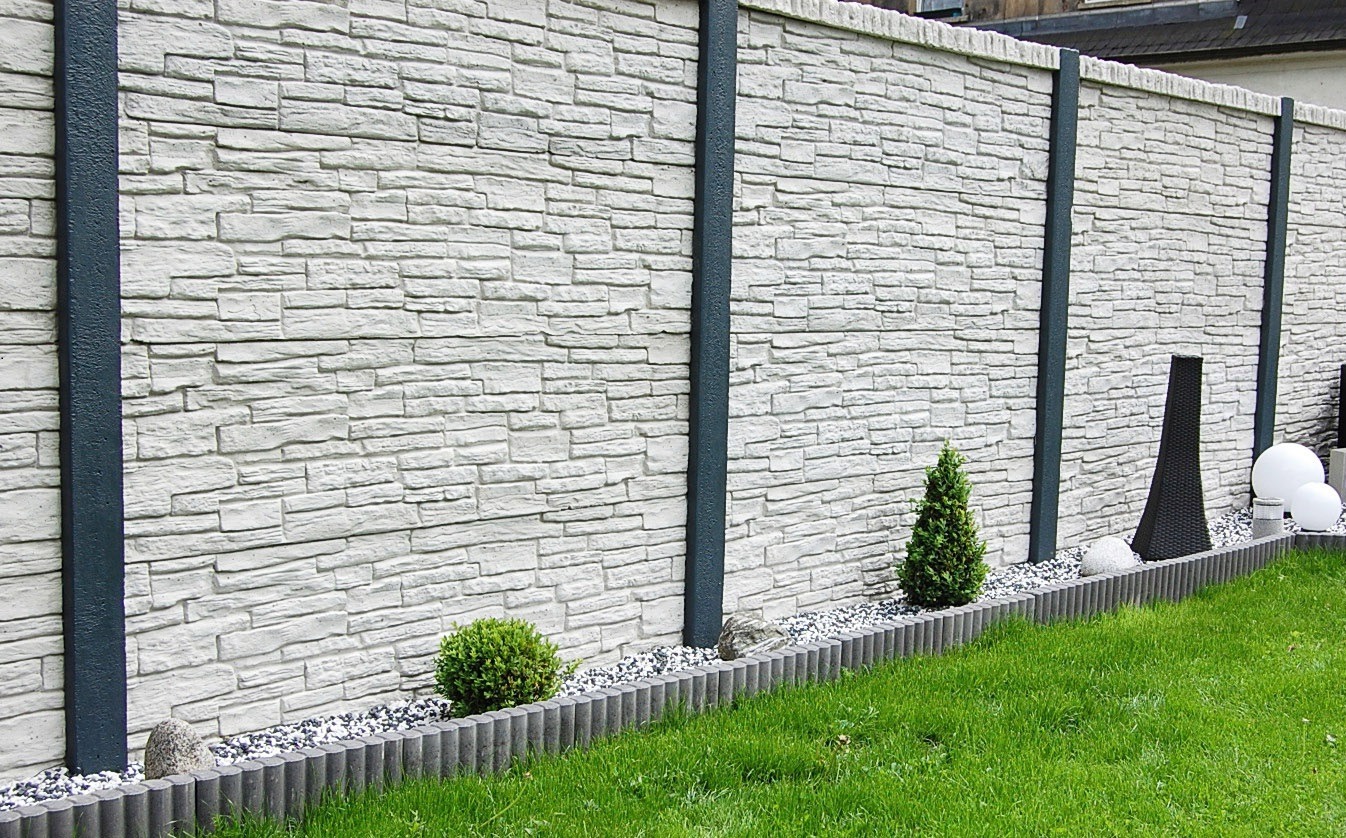 Szary, betonowy płot z imitacją kamienia, ciemne słupki, oddzielony od trawnika obrzeżem z kostki i białym żwirem, dekoracyjne krzewy i kule.