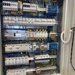 Homel Łukasz Dudziak - Otwarta szafa elektryczna z licznymi zabezpieczeniami, przewodami oznaczonymi żółtymi etykietami i elementami sterującymi, uporządkowany układ w szarej obudowie z widocznym uziemieniem.