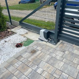 Homel Łukasz Dudziak - Szara brama przesuwna z zamontowanym napędem automatycznym, widoczna na tle posesji z podjazdem z kostki brukowej i ogrodzeniem z siatki.