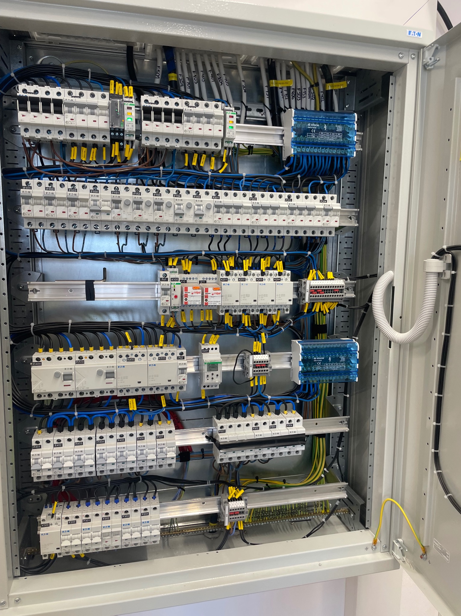 Otwarta szafa elektryczna z licznymi zabezpieczeniami, przewodami oznaczonymi żółtymi etykietami i elementami sterującymi, uporządkowany układ w szarej obudowie z widocznym uziemieniem.