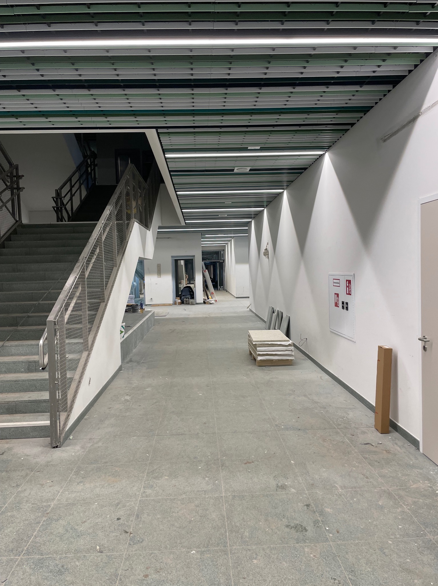 Wnętrze budynku w trakcie wykańczania: widoczne schody z metalową balustradą, korytarz z płytkami na podłodze, białe ściany z instalacją przeciwpożarową i strop z oświetleniem liniowym. W tle...