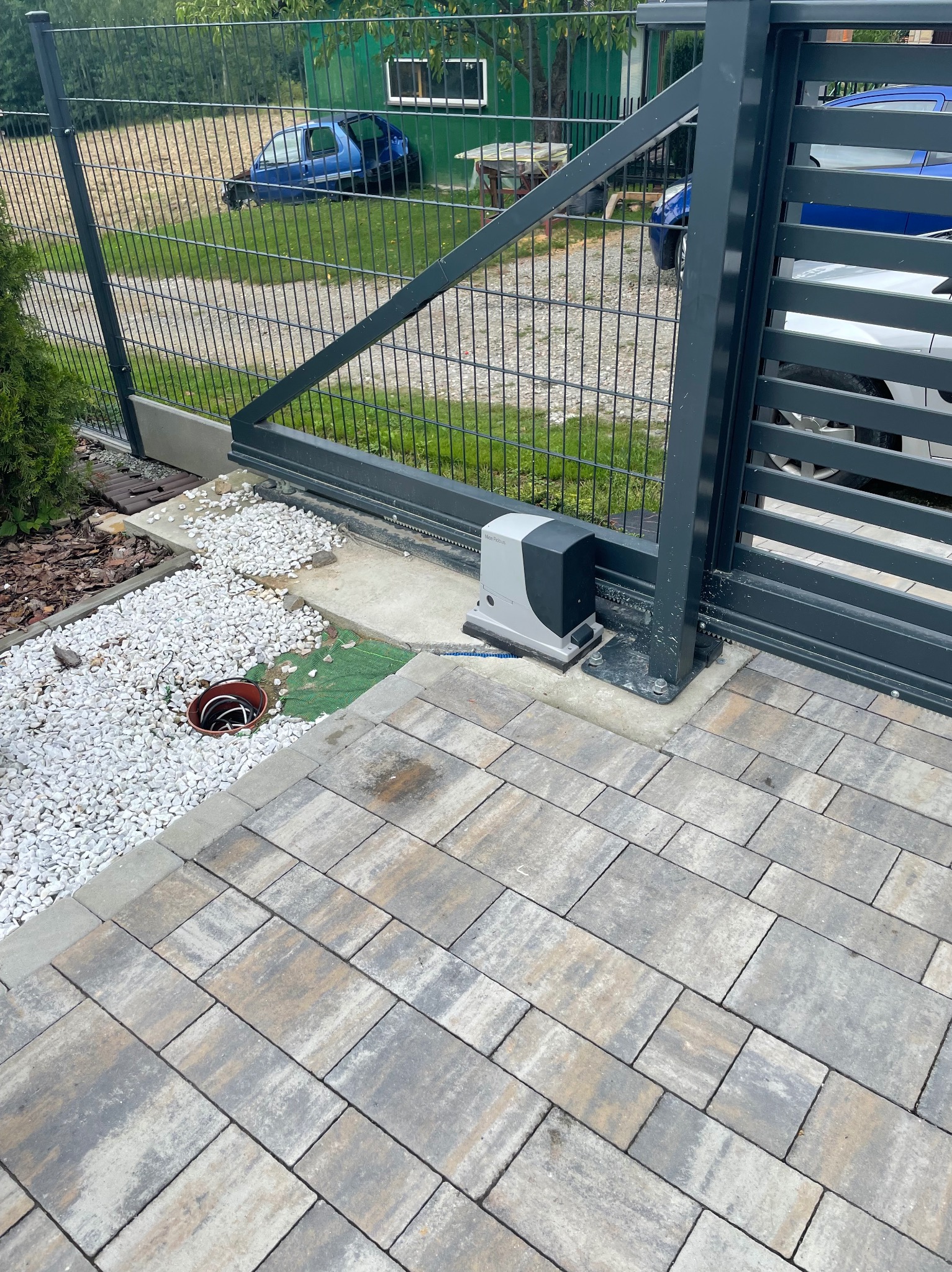 Szara brama przesuwna z zamontowanym napędem automatycznym, widoczna na tle posesji z podjazdem z kostki brukowej i ogrodzeniem z siatki.