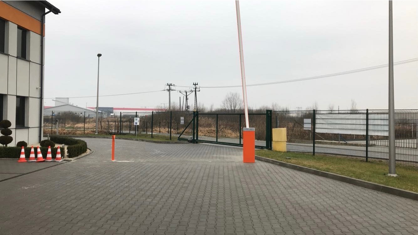Automatyczna barierka podniesiona przy wjeździe na teren przemysłowy, widoczne słupy energetyczne w tle, oraz rząd pomarańczowych pachołków przy budynku.