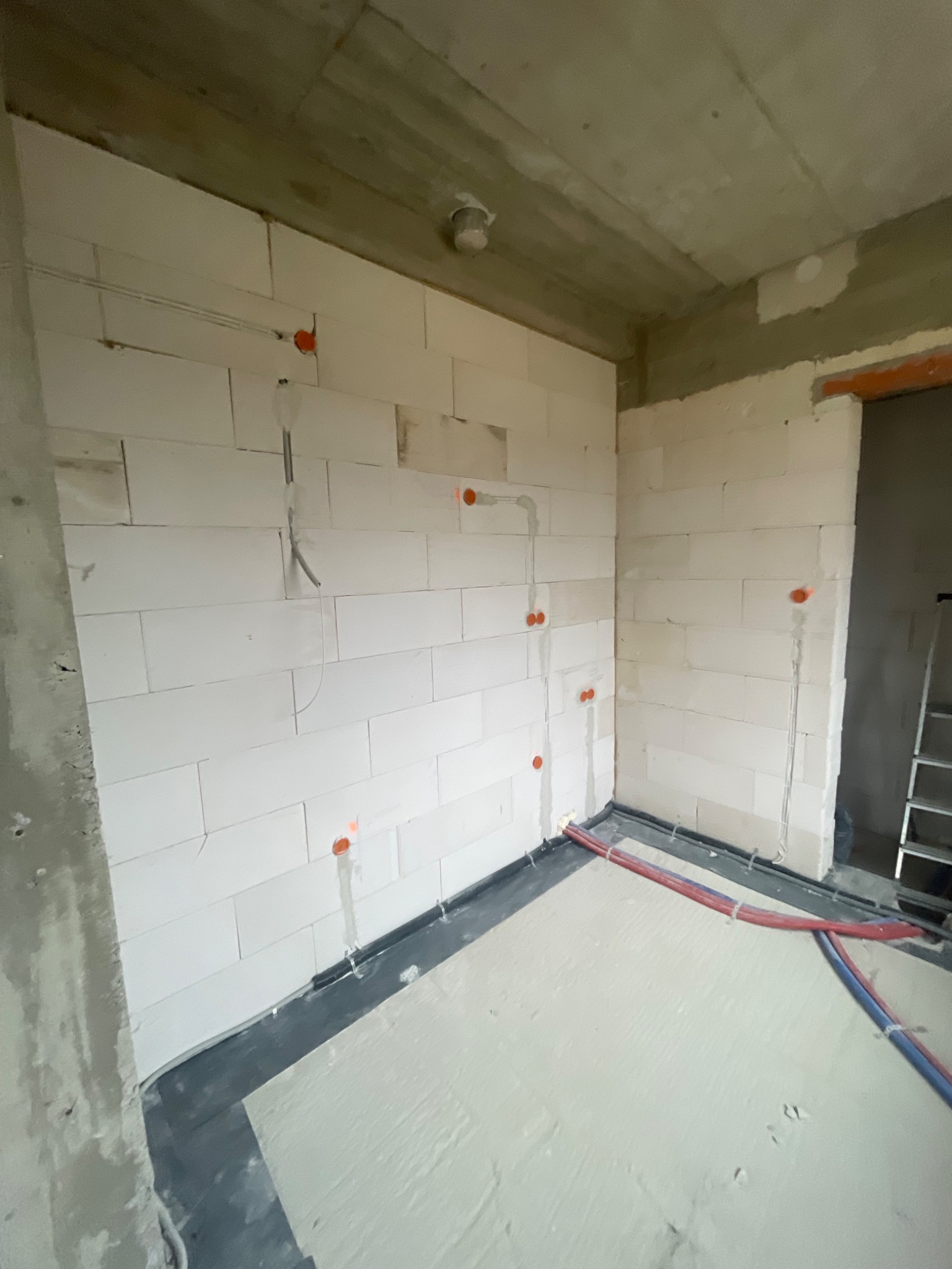 Wnętrze w trakcie remontu z widocznymi przewodami elektrycznymi wychodzącymi ze ścian z bloczków, oraz rurami grzewczymi przy podłodze.