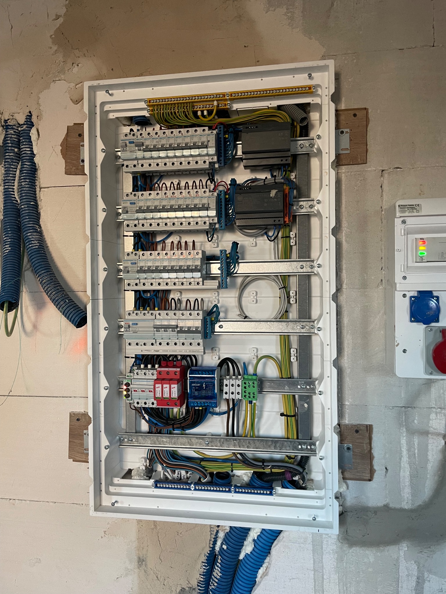Otwarta, biała skrzynka rozdzielcza z bezpiecznikami, przewodami i innymi elementami, zamontowana na surowej ścianie. Widoczne niebieskie rury karbowane doprowadzające kable. Obok zamontowana...