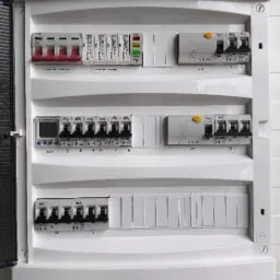 Otwarta skrzynka elektryczna firmy Schrack Technik z bezpiecznikami, wyłącznikami różnicowoprądowymi i licznikiem energii, widoczne oznaczenia na poszczególnych elementach.