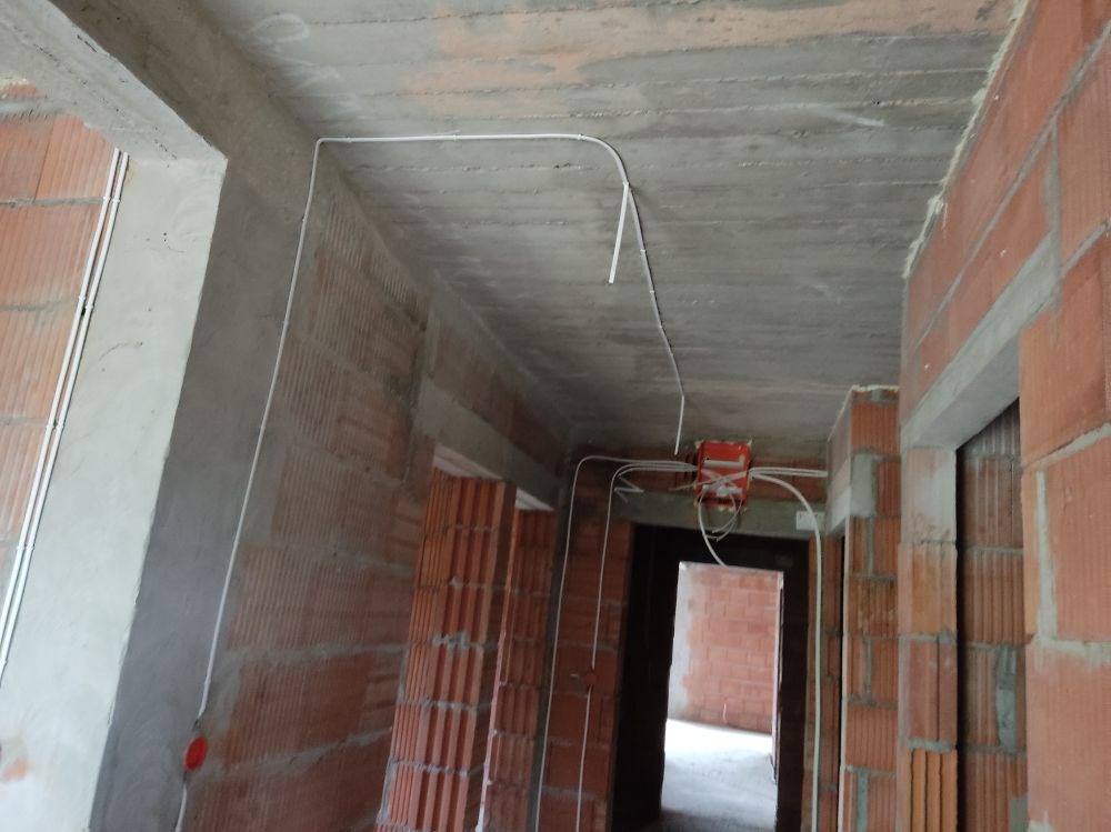 Wnętrze w stanie surowym: widoczne przewody elektryczne prowadzone po betonie sufitowym i ścianach z czerwonej cegły, puszka elektryczna zamontowana nad otworem drzwiowym.