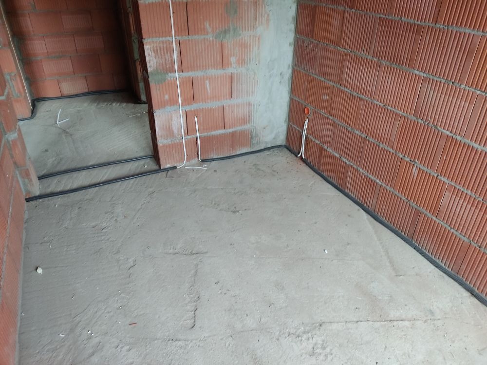 Surowy stan wnętrza z widocznymi przewodami w listwach przypodłogowych i kablami wystającymi ze ściany, ściany z czerwonej cegły dziurawki, wylewka podłogowa w trakcie schnięcia.