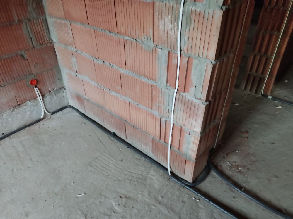 Wewnętrzna instalacja elektryczna w nowo budowanym domu, przewody prowadzone wzdłuż ściany z czerwonej cegły, widoczna puszka elektryczna.