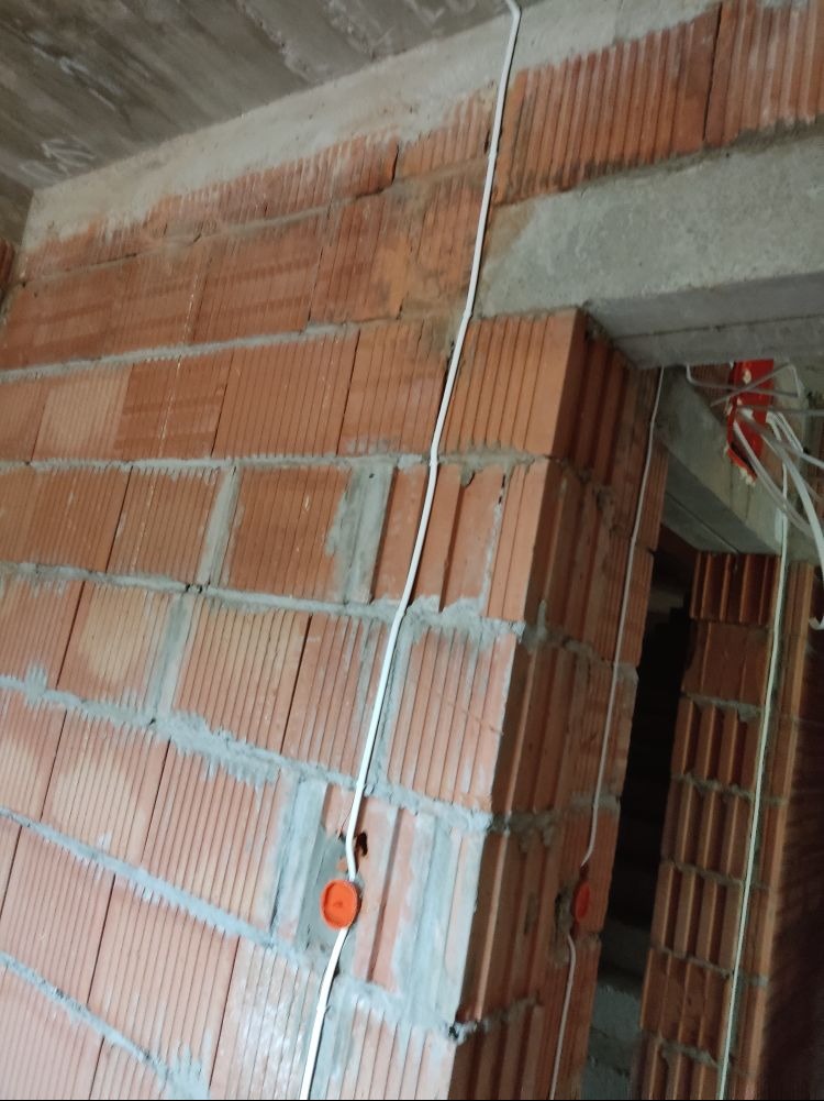 Biały przewód elektryczny poprowadzony po czerwonej cegle dziurawce, widoczne puszki instalacyjne w kolorze pomarańczowym, surowy stan wykończenia.