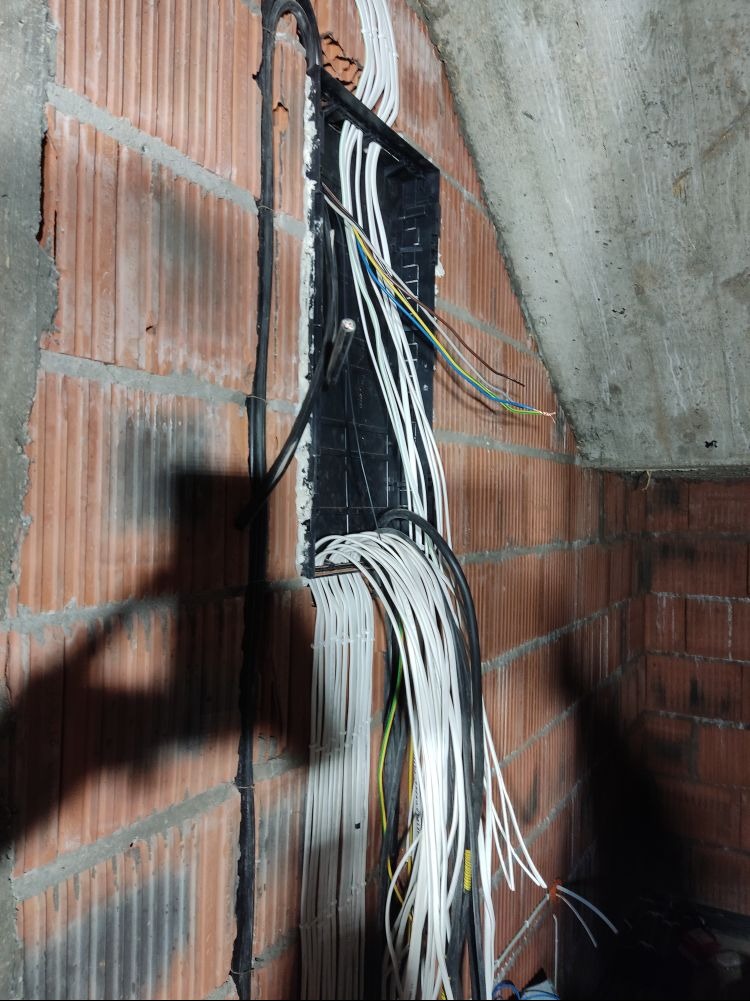 Ściana z cegły z wyciętym otworem, przez który przechodzą liczne kable elektryczne, zarówno białe, jak i czarne oraz kolorowe, prowadzone w peszlu, widoczne elementy instalacji elektrycznej...