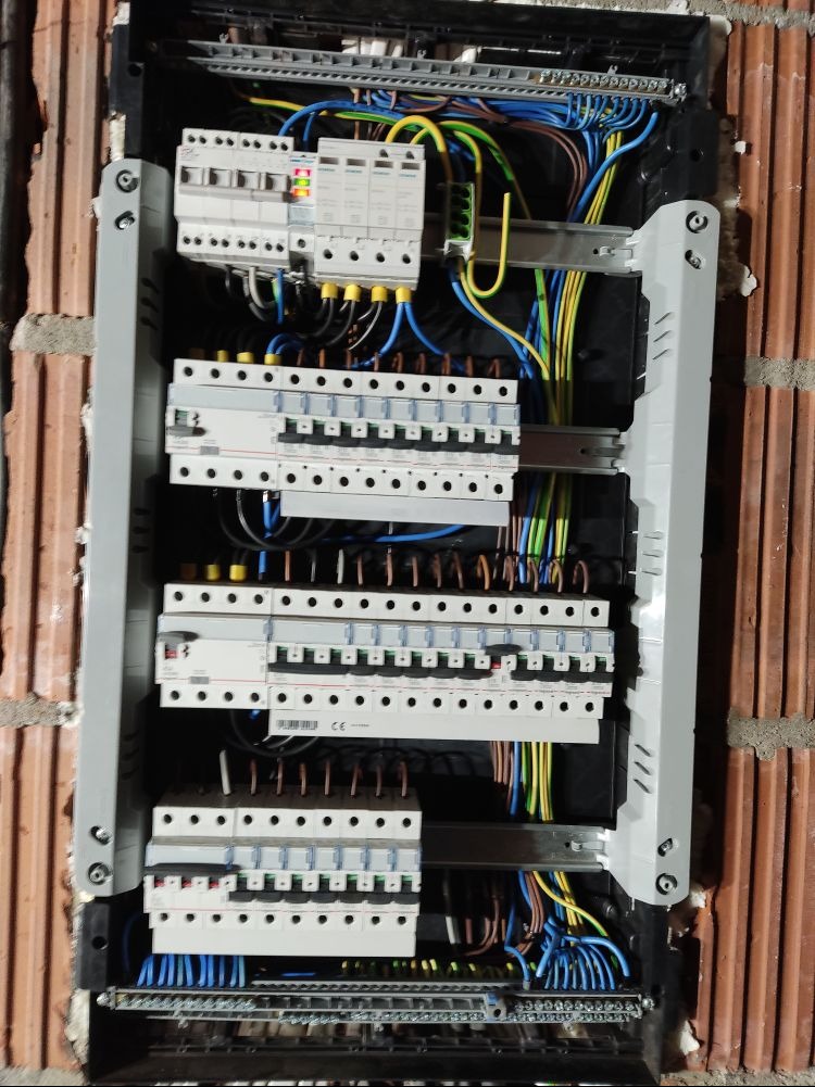 Rozdzielnica elektryczna z licznymi bezpiecznikami, przewodami w kolorach niebieskim, żółto-zielonym i brązowym, zamontowana w ścianie z czerwonej cegły.