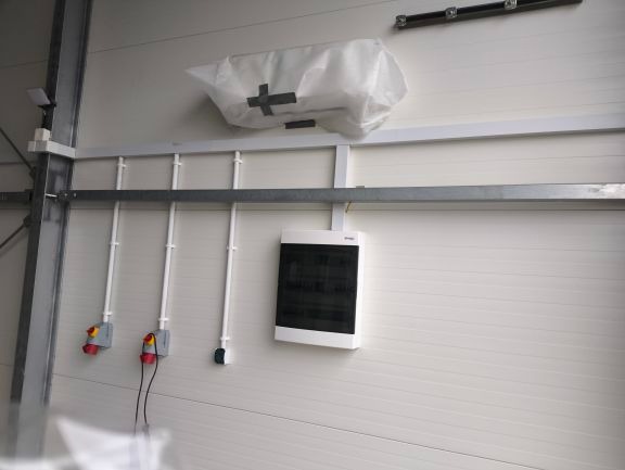 Biała ściana z zamontowaną skrzynką elektryczną, trzema pionowymi rurkami z przewodami, gniazdkami i szarą metalową szyną. Na górze widoczny biały worek zabezpieczony taśmą.