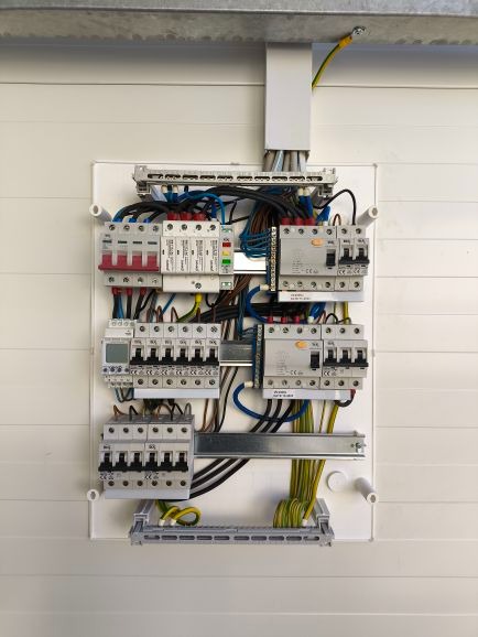 Rozdzielnica elektryczna z widocznymi bezpiecznikami, przewodami i elementami montażowymi w białej obudowie, zamontowana na ścianie z poziomymi panelami.