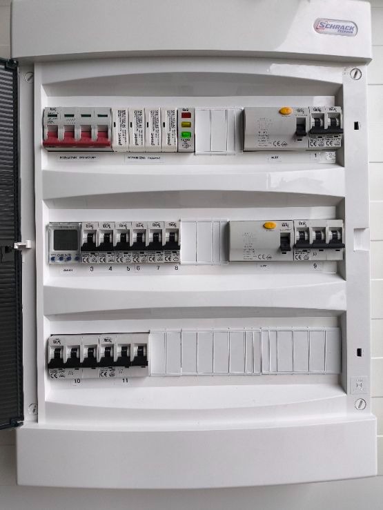 Otwarta skrzynka elektryczna firmy Schrack Technik z bezpiecznikami, wyłącznikami różnicowoprądowymi i licznikiem energii, widoczne oznaczenia na poszczególnych elementach.