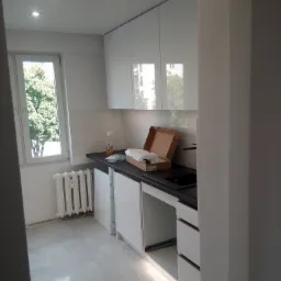 Nowoczesna kuchnia w trakcie montażu, białe szafki z połyskiem, czarny blat, widoczny karton z akcesoriami i środki czystości na blacie, płytki podłogowe, oświetlenie podszafkowe i grzejnik pod oknem.