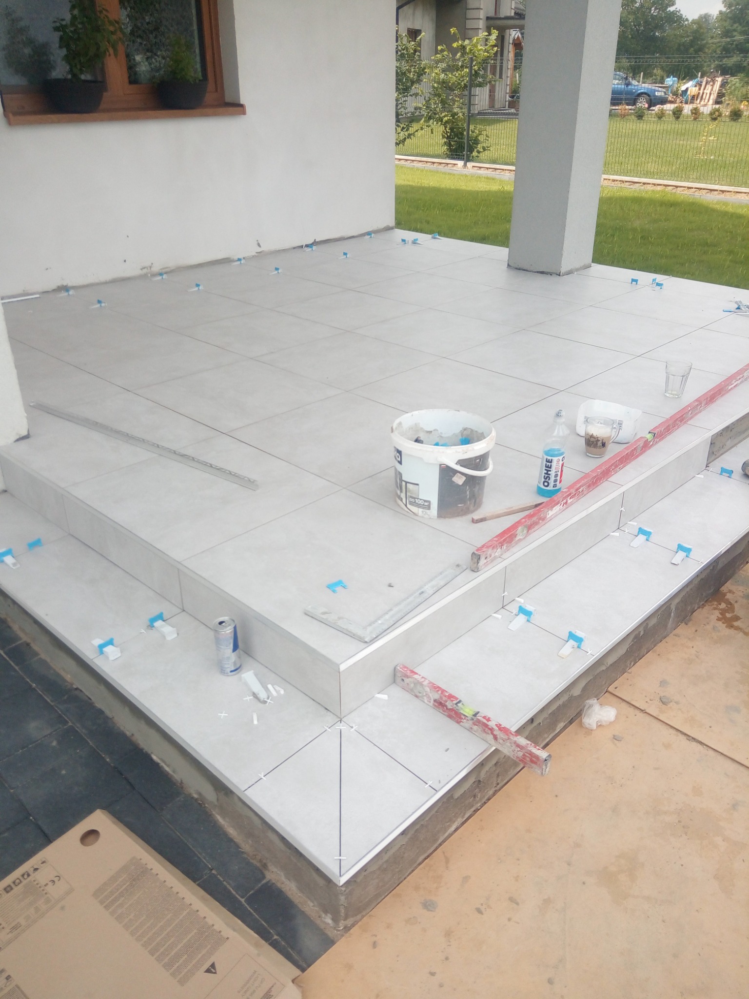 Układanie jasnych, kwadratowych płytek na schodach wejściowych domu, widoczne narzędzia: poziomica, puszka, wiadro z klejem, krzyżyki dystansowe, napój energetyczny.