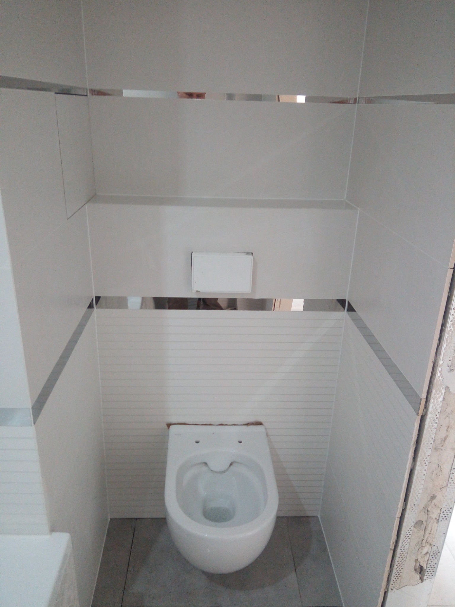 Zamontowana toaleta wisząca w małej łazience wykończonej jasnymi płytkami z poziomymi paskami, z wąskim srebrnym akcentem dekoracyjnym, widoczny fragment futryny drzwi w trakcie montażu.