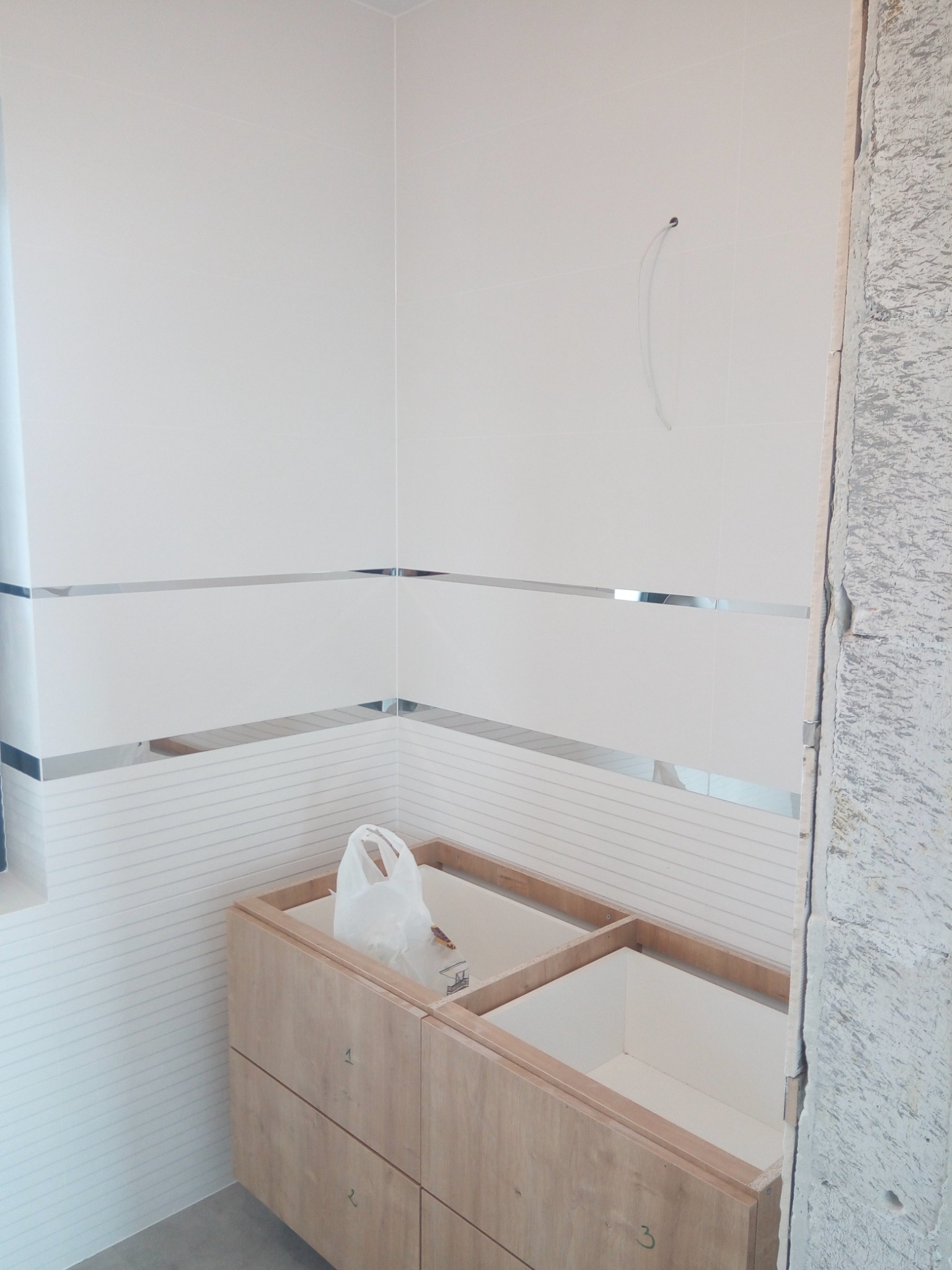 Łazienka w trakcie wykańczania: białe płytki z poziomymi srebrnymi paskami, drewniana szafka pod umywalkę bez blatu, surowy fragment ściany po prawej.