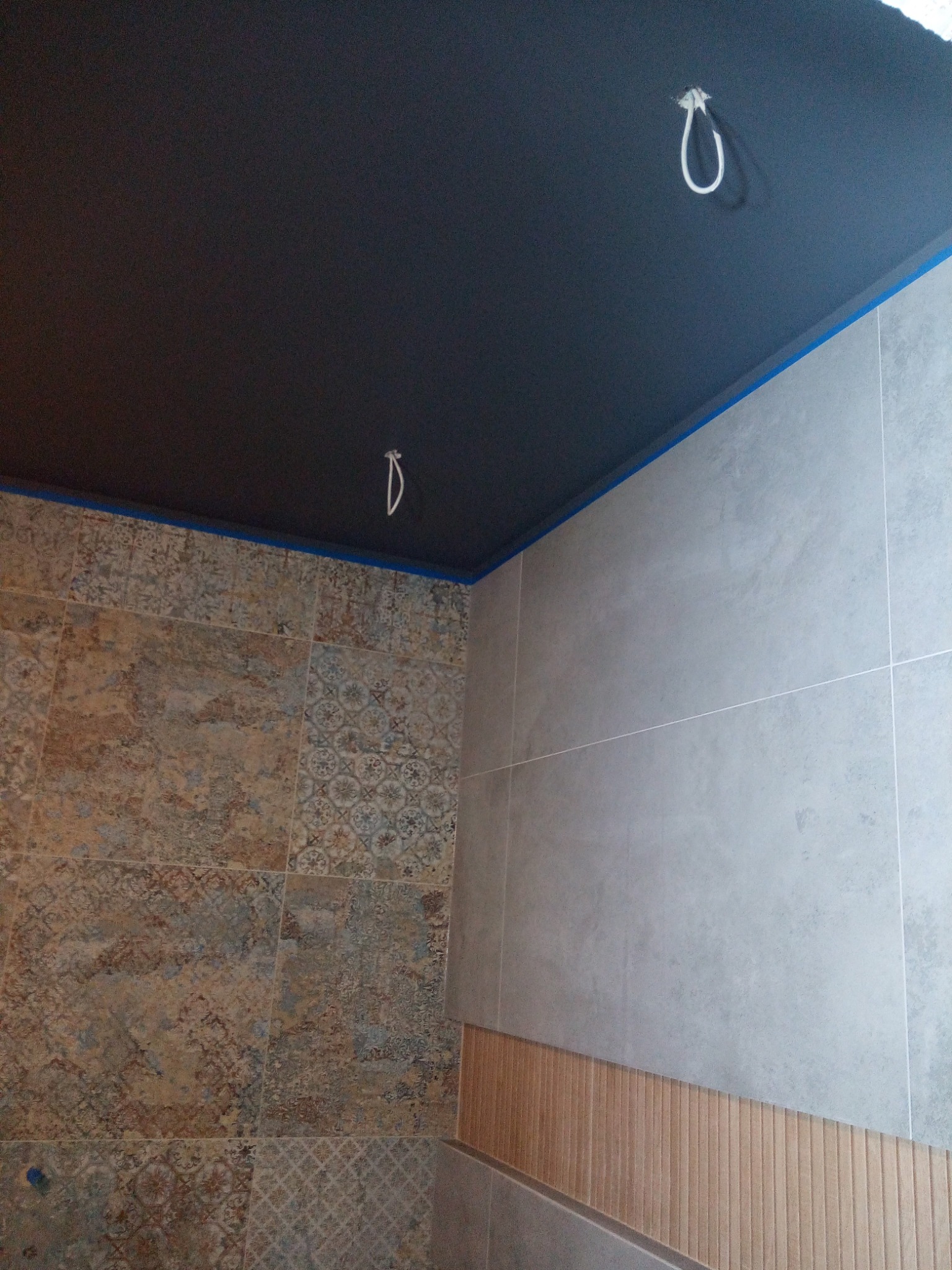 Ciemny, matowy sufit z wystającymi kablami oświetleniowymi w nowocześnie wykończonej łazience z mozaikowymi i gładkimi, szarymi płytkami, zabezpieczony niebieską taśmą malarską.