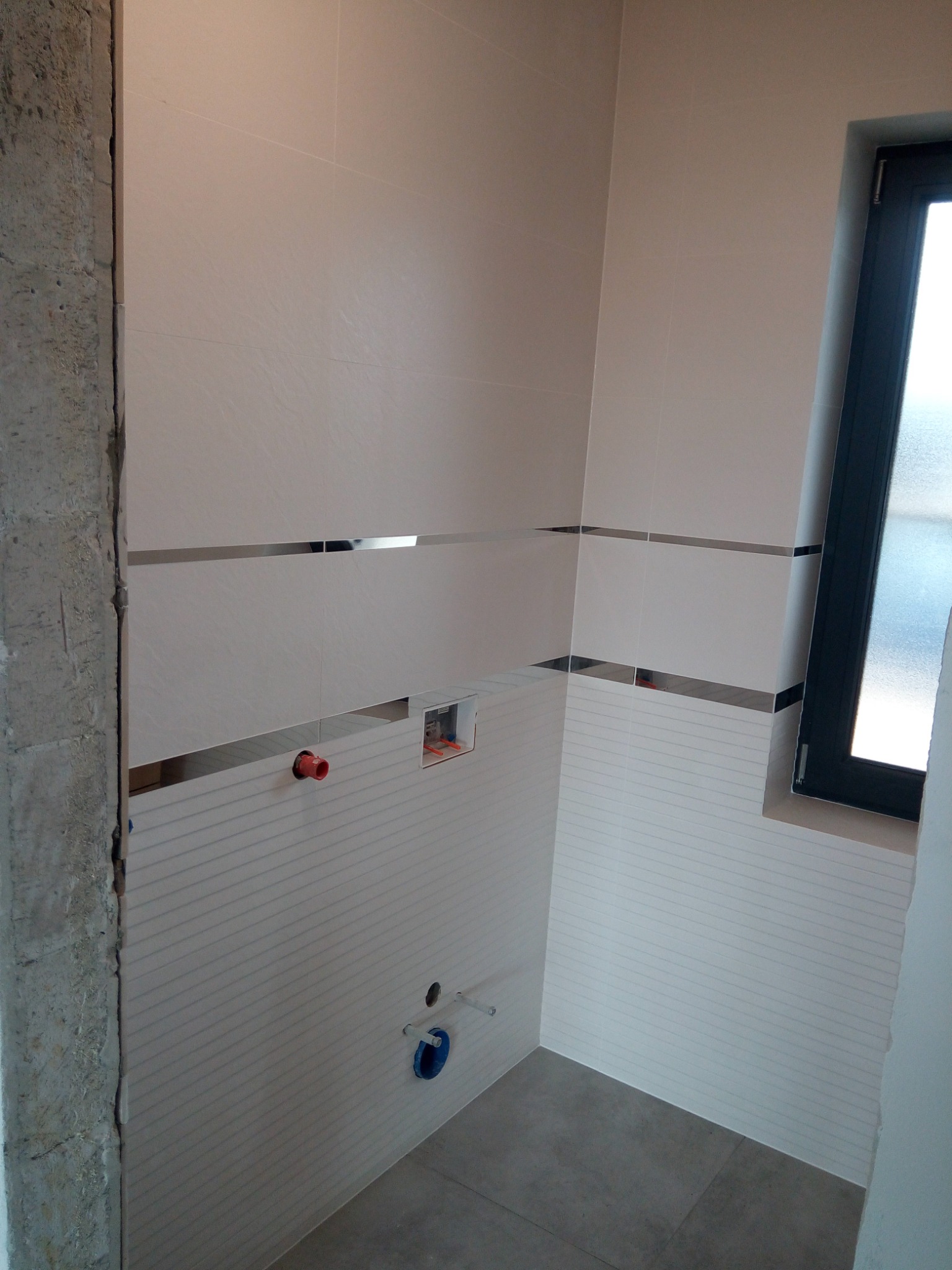 Ściany łazienki w trakcie wykańczania, z widocznymi białymi płytkami z poziomymi żłobieniami, oddzielonymi pasami z metalowymi listwami, niezabudowany narożnik z surowego betonu, okno z czarną ramą.